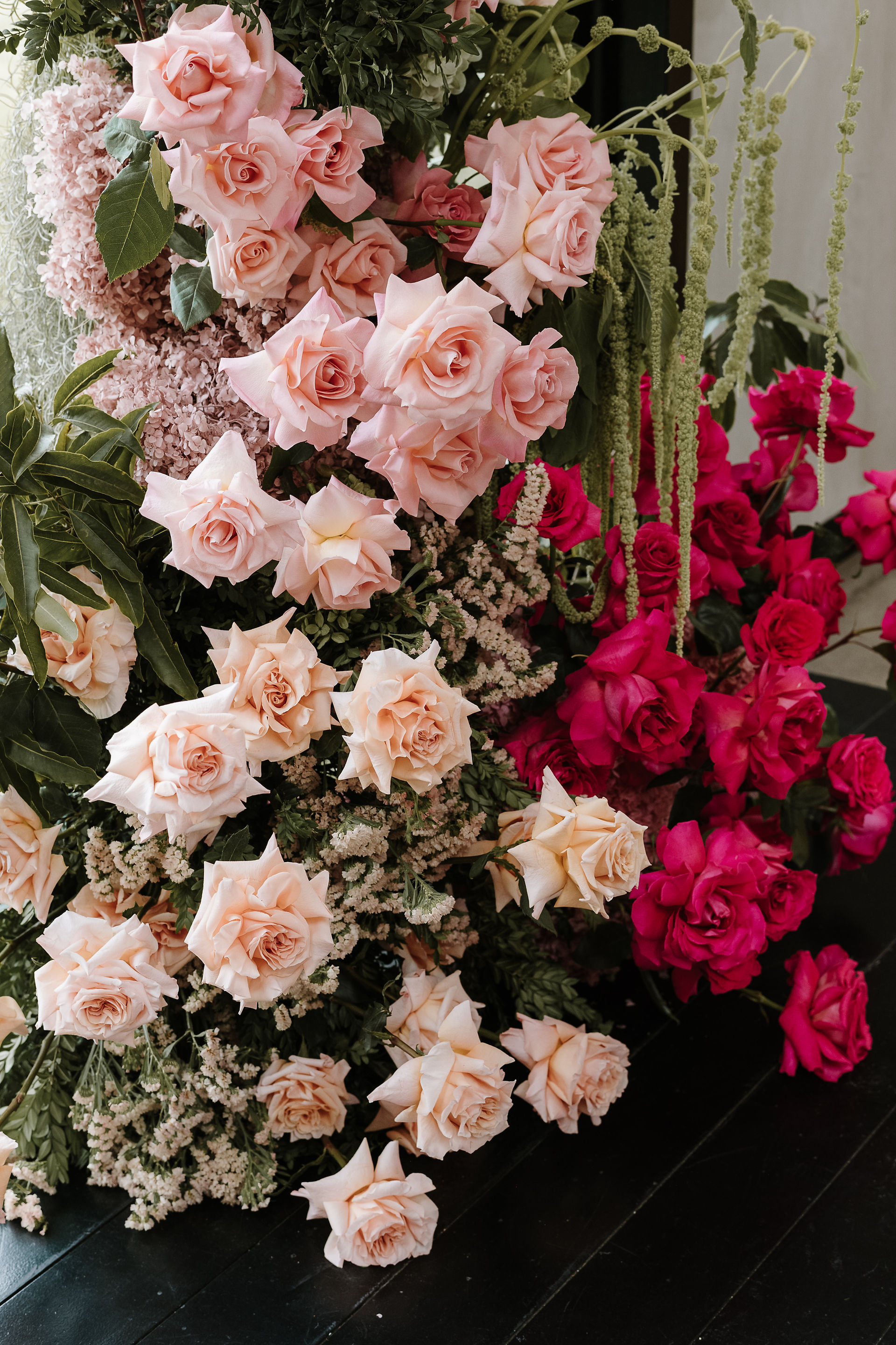wo asymmetrical floral pillars featuring vibrant red, pink, and peach roses with green amaranthus stand before a large grid window overlooking the Zonzo Estate vineyard.