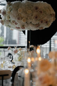 An elegant wedding reception setup featuring round tables with white tablecloths, modern white and black cushioned chairs, and tall, voluminous floral centerpieces made of white and light pink roses. The tables are meticulously set with crystal glassware, lit taper candles, and small gold perfume bottles as favors. In the background, similar tables stretch into a dimly lit room with black draped windows and a layered fabric ceiling installation.