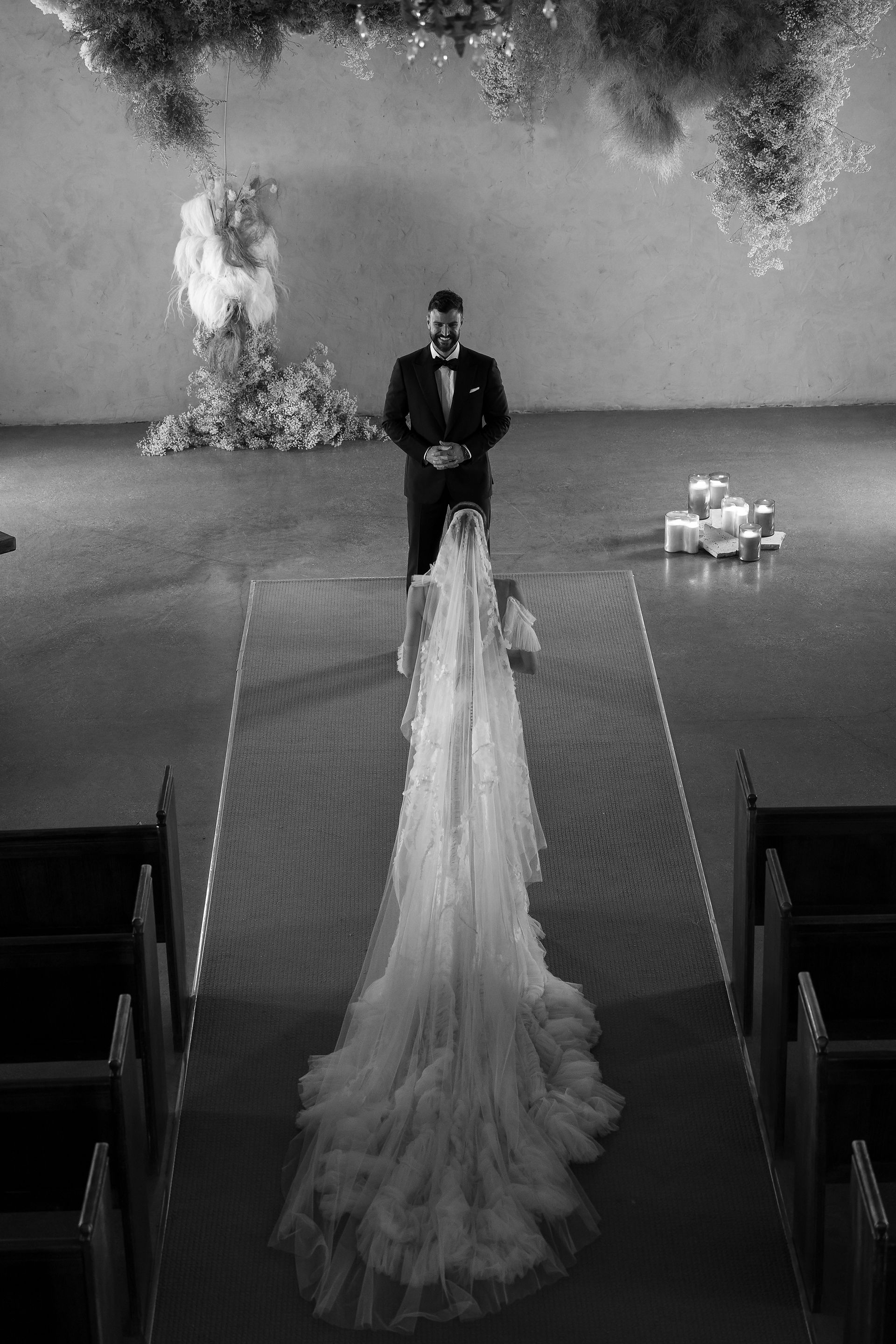 A bride and groom stand facing each other in the luxurious Stones of the Yarra Valley venue, adorned with elegant floral arrangements, candles, and a large stained glass window. The bride wears a flowing veil and holds a bouquet; the groom is in a black tuxedo.