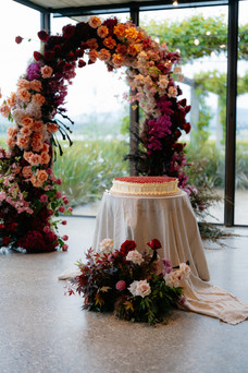 A wide, single-tier wedding cake topped with red berries on a linen-draped table. It is framed by a large, vibrant, asymmetrical floral arch featuring burgundy, magenta, and peach flowers, set indoors in front of large glass windows overlooking greenery.