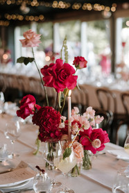 Close-up of a table setting featuring a cocktail with a mint garnish next to a vibrant floral arrangement of bright red roses, sweet peas, and a single white taper candle.