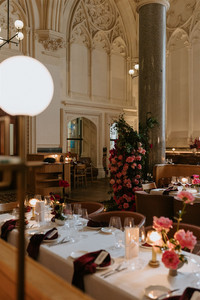 A warm dining room view through soft globe lighting, showing tables set with white linens, pink flowers, and a large rose installation around a stone pillar.