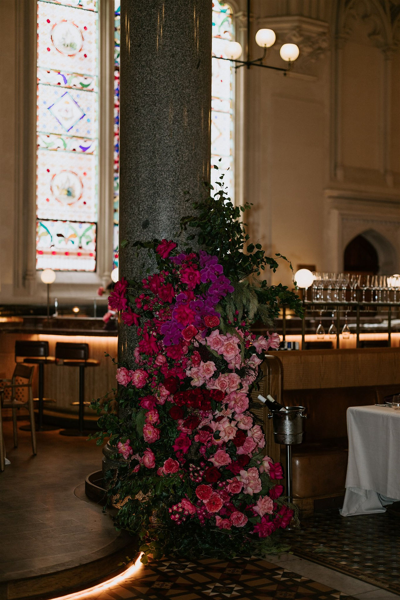 A towering floral pillar of pink, red, and purple blooms stands against a stone column, with stained-glass windows and a bar area in the background.