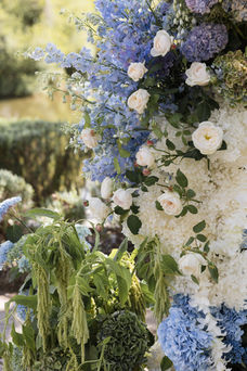 A macro view of floral arrangement details, highlighting light blue delphiniums, scattered white garden roses, lush white hydrangeas, and textured green hanging amaranthus.
