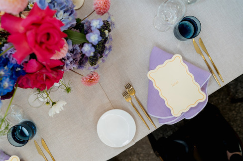 A beautifully set table illuminated by sunlight, featuring a vibrant floral centerpiece with pink, orange, white, and purple flowers. There are also tall candles, wine glasses, and a bread basket. Perfect for a wedding at Stones of the Yarra Valley, the table is elegantly arranged with plates and cutlery.