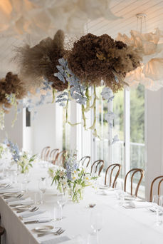 A long banquet table with white linens, bentwood chairs, and blue and white floral centerpieces, set beneath large hanging floral installations of dried grasses and cascading flowers in a bright, window-lined room