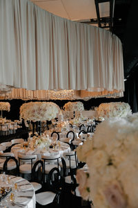An elegant wedding reception setup featuring round tables with white tablecloths, modern white and black cushioned chairs, and tall, voluminous floral centerpieces made of white and light pink roses. The tables are meticulously set with crystal glassware, lit taper candles, and small gold perfume bottles as favors. In the background, similar tables stretch into a dimly lit room with black draped windows and a layered fabric ceiling installation.