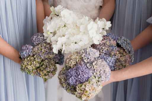 Close-up of a central white orchid bridal bouquet surrounded by three bridesmaids' bouquets made of rustic blue, purple, and green hydrangeas against light blue pleated dresses.