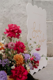 A vibrant floral arrangement featuring a variety of flowers. Prominent blooms include pink peonies, red roses, and clusters of purple and blue hydrangeas, complemented by delicate white and yellow daisies. The elevated design sits against the welcome sign