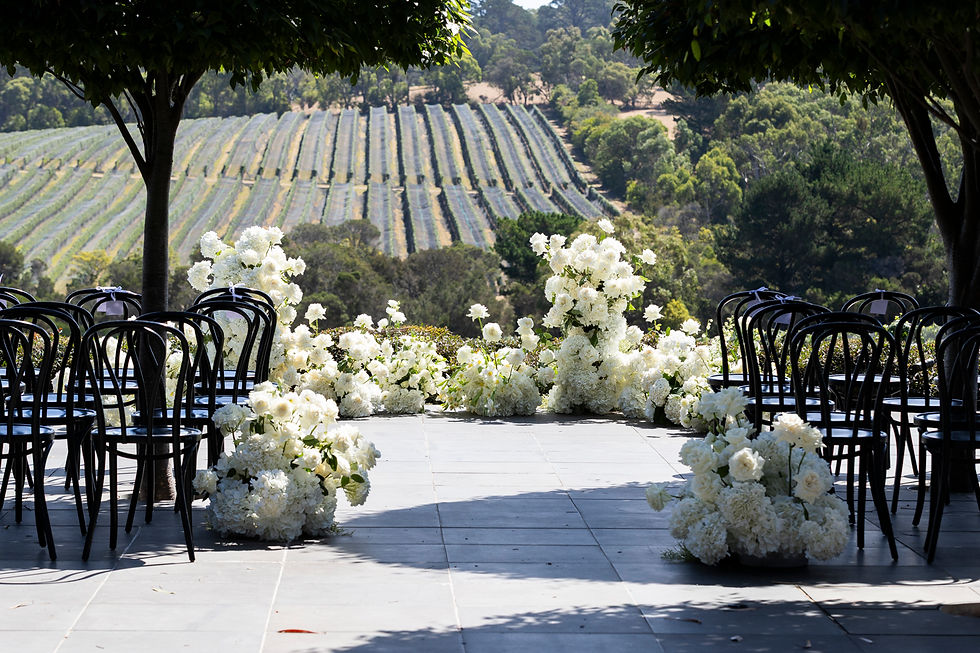 An outdoor wedding ceremony at Jackalope featuring black bentwood chairs on a stone terrace. Lush white floral arrangements frame a breathtaking view of a rolling, netted vineyard landscape.