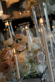 An elegant wedding reception setup featuring round tables with white tablecloths, modern white and black cushioned chairs, and tall, voluminous floral centerpieces made of white and light pink roses. The tables are meticulously set with crystal glassware, lit taper candles, and small gold perfume bottles as favors. In the background, similar tables stretch into a dimly lit room with black draped windows and a layered fabric ceiling installation.