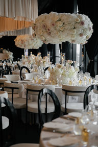 An elegant wedding reception setup featuring round tables with white tablecloths, modern white and black cushioned chairs, and tall, voluminous floral centerpieces made of white and light pink roses. The tables are meticulously set with crystal glassware, lit taper candles, and small gold perfume bottles as favors. In the background, similar tables stretch into a dimly lit room with black draped windows and a layered fabric ceiling installation.
