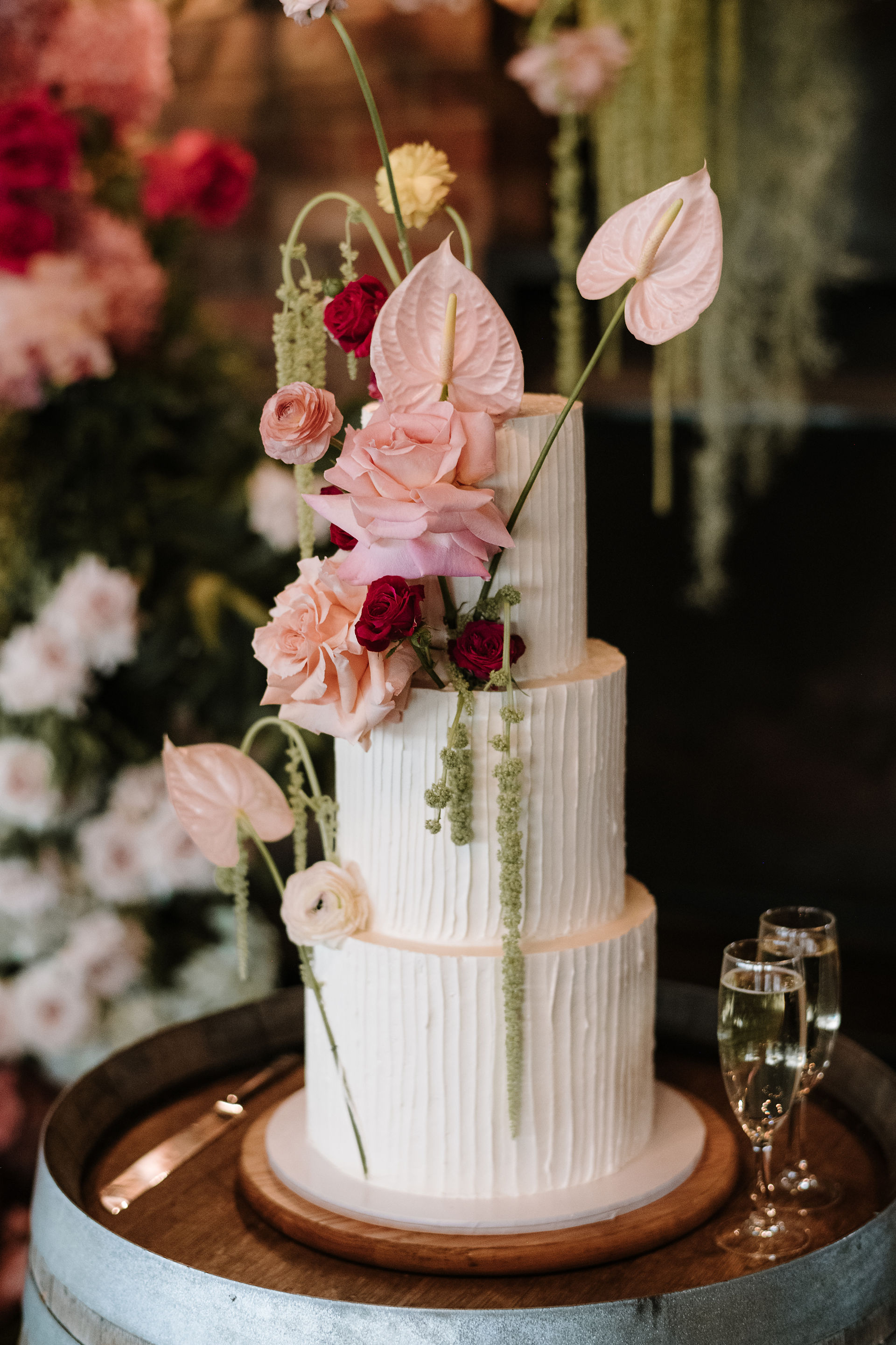 A vertical close-up of the wedding cake decorated with fresh pink roses, red spray roses, and long strands of green amaranthus, set against a dark, moody background.