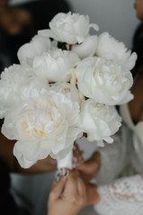 A close-up shot focusing on a bridal bouquet made entirely of large, fluffy white peonies. The hands of the bride, showcasing manicured nails and a glimpse of her white lace sleeve, hold the stems tightly wrapped in a white ribbon.