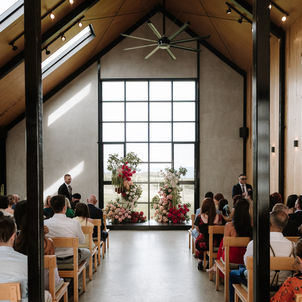 A wedding ceremony at Zonzo Estate. Guests sit in wooden pews facing a large window and vibrant pink, red, and white amaranthus floral pillars during a bright indoor service.