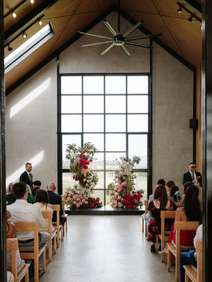 A wedding ceremony at Zonzo Estate. Guests sit in wooden pews facing a large window and vibrant pink, red, and white amaranthus floral pillars during a bright indoor service.