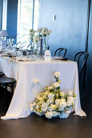 Elegant indoor reception at Jackalope featuring long tables with white linens, black-edged napkins, and bentwood chairs. Minimalist glass vases hold white roses and calla lilies beneath pleated table lamps.