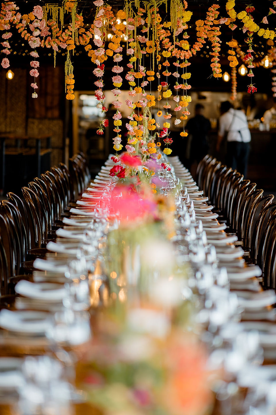 Elegant wedding table setting featuring a tall wildflower centerpiece with pink zinnias and red roses, flanked by sparkling glassware on a polished wood surface.