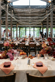 A close-up detail shot of an elegant wedding table setting at Stones of the Yarra Valley. The arrangement features a dark, textured dinner plate and gold cutlery resting on a crisp white tablecloth. A softly glowing candle in a clear glass holder provides warm light, highlighting the moody, sophisticated aesthetic of the reception.