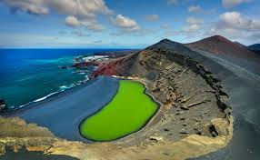 A close-up of the Charco Verde (Green Lagoon) in Lanzarote. A small, intensely green pool of water is separated from the blue ocean waves by a strip of black volcanic sand. Behind the lagoon, the dramatic, eroded amphitheater of a volcanic crater shows reddish-brown and orange rock layers.