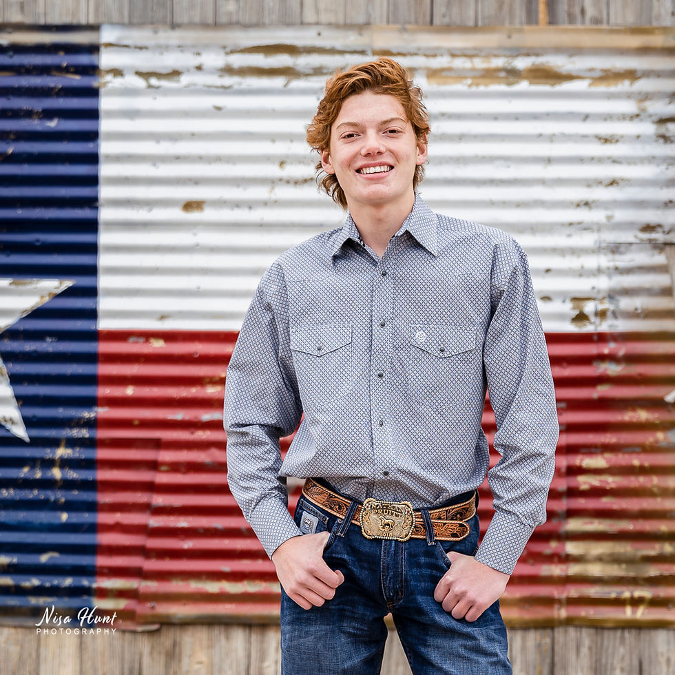 Smiling man in a patterned shirt and jeans stands in front of a corrugated metal wall painted like a faded Texas flag, exuding a relaxed vibe.