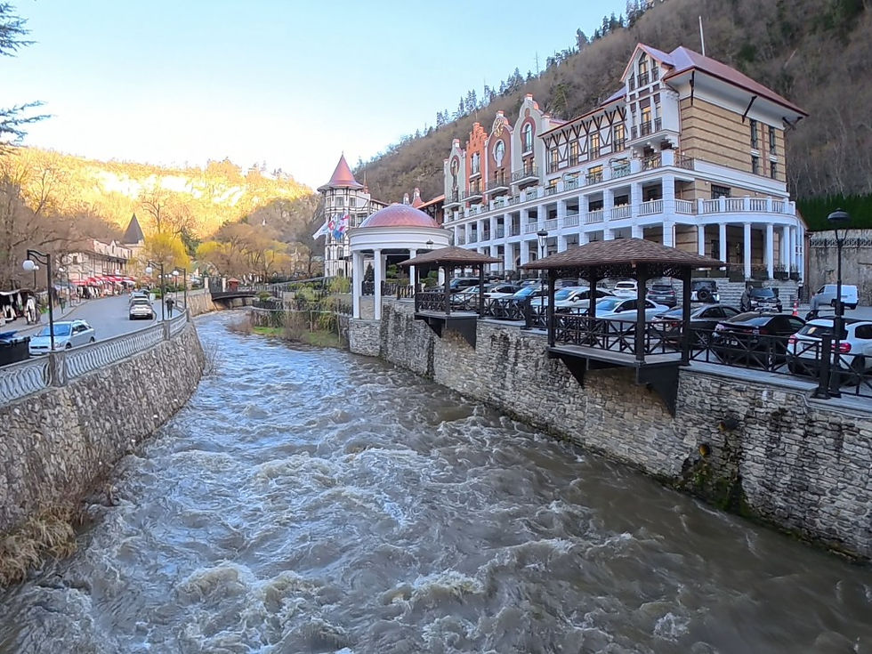 River flows alongside a stone wall, flanked by a large hotel with red roofs and cars parked outside. Trees and hills under a clear sky.