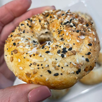 Hand holding a golden-brown bagel topped with sesame seeds and spices. Pink nails visible. Background has a blurred out breakfast setting.