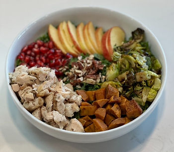 Bowl of salad with chicken, sweet potatoes, Brussels sprouts, pecans, apples, and pomegranate arils, on a white table. Colorful and fresh.