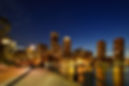 A nighttime view of the Boston city skyline  and in the foreground a walking path lit by a streetlamp runs along the water