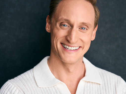 Smiling person in a white knit shirt, arms crossed. Dark background enhances the bright outfit and cheerful expression.