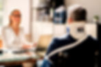 A man in a wheelchair with a neck brace sits at a desk across from a woman with a laptop, in a bright office setting.
