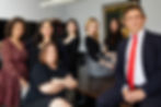 Seven people in formal attire pose confidently in an office. A poster with text "CACHOU LAJAUNIE" is on the wall behind them.