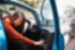 Woman in orange sweater holds head in pain inside a blue car after an accident. Smoke is visible outside. Mood is tense and concerned.