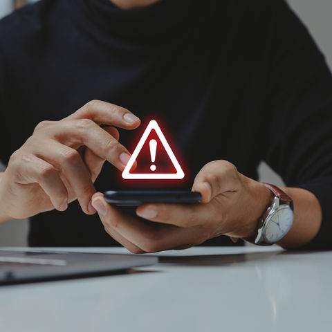 Person in dark clothing holds a smartphone showing a glowing red warning symbol. Laptop partially visible on a white table.
