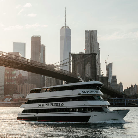 Boat "Skyline Princess" sails near Brooklyn Bridge against a backdrop of New York City skyscrapers at sunset, with calm water reflecting light.