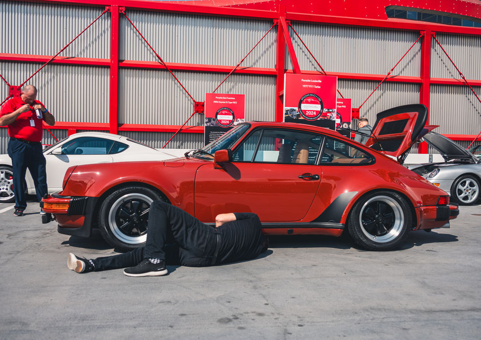 Man in black clothes doing work under a red vintage sports car sitting in a parking lot on a bright sunny day.