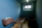 A sparse prison cell with a wooden bed frame, desk with papers, single window, and blue walls, creating a stark, somber atmosphere.