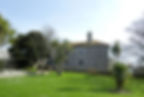 Stone building with a rustic roof set in a lush green garden. Palm trees and sparse leafless trees surround it. Clear blue sky above.