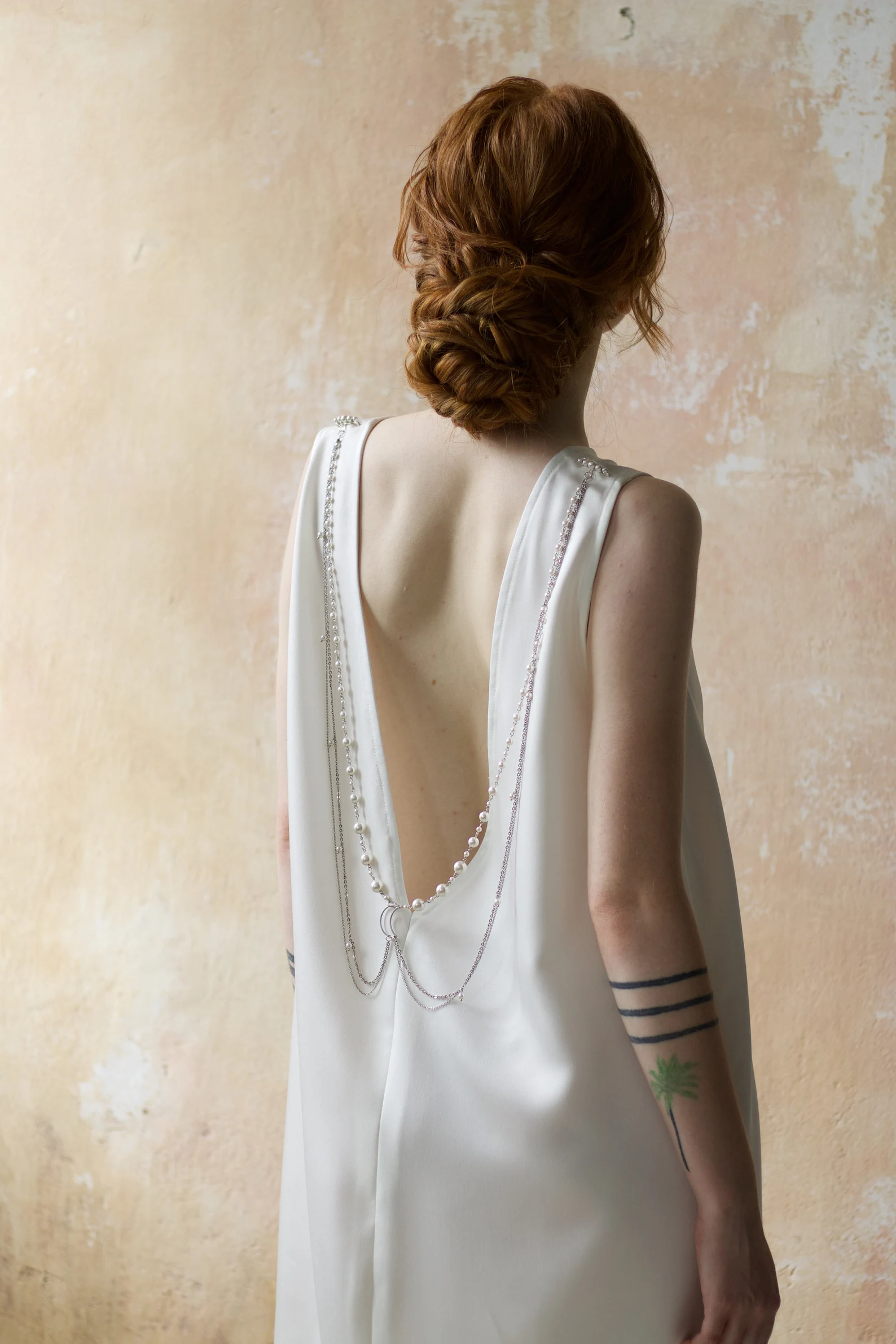 Back view of a young woman with dark red hair wearing a white dress and silver silver body chain with pearls