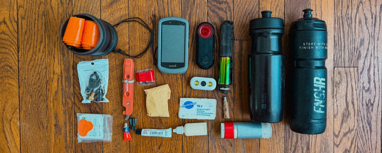 Outdoor gear and first-aid items laid out on a wooden floor: water bottles, GPS, flashlight, pouches, and supplies.