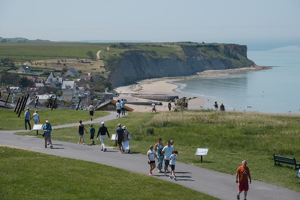 Tourists walk a grassy cliff path above a sandy beach and calm sea, with seaside houses and a 1944 marker.