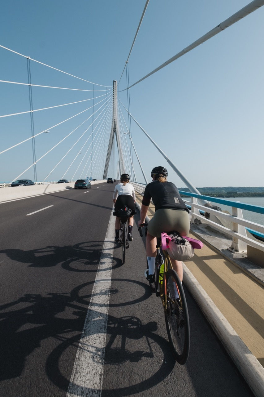 Two cyclists ride across a cable-stayed bridge under a clear blue sky, with car traffic and water below.