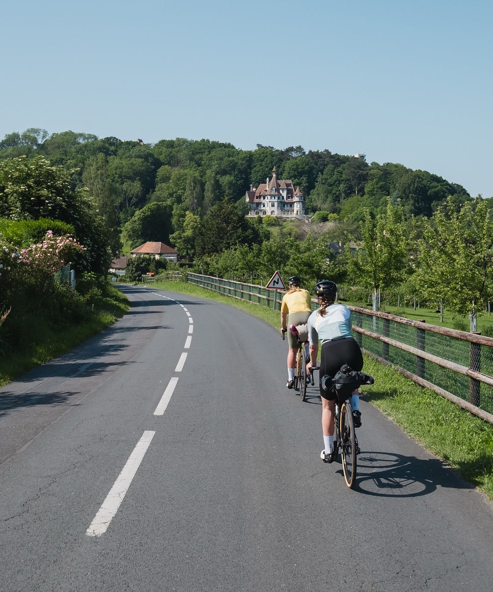 Two cyclists ride along a curving country road toward a hilltop castle, with green trees, fences, and a sunny blue sky.