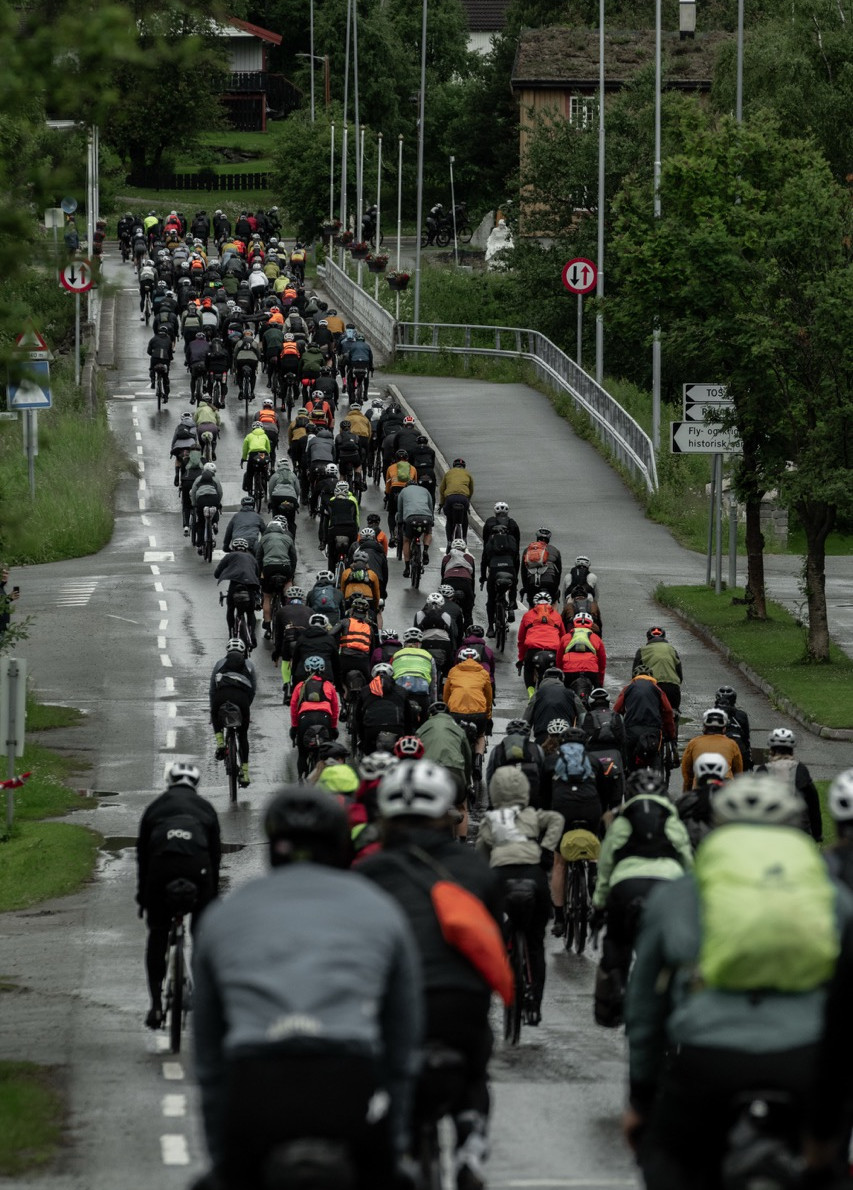 Large group of cyclists rides uphill on a wet road in a green village, with road signs and a rainy, overcast mood.