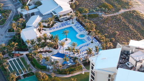 Aerial view of a large commercial pool complex with rectangular pool, spa, palm trees, lounge chairs, and nearby resort buildings near a beachside dune.