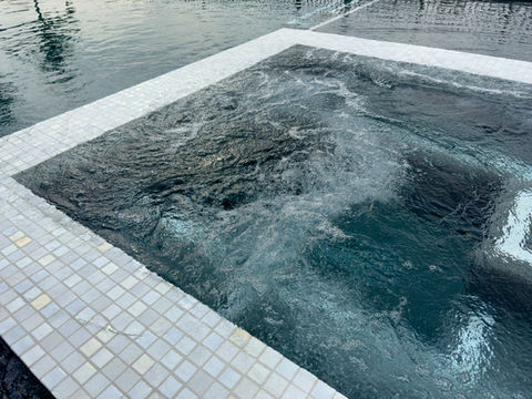 A modern spa with white mosaic tiles and bubbling jets, seamlessly integrated into a larger pool with dark water.