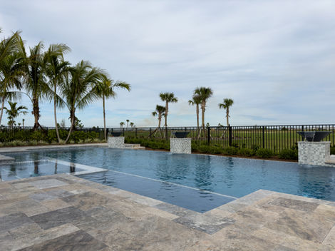 A wide pool with a decorative tiled path, fire bowls on plinths, and palm trees in the background, set against a cloudy sky and a distant fence line.