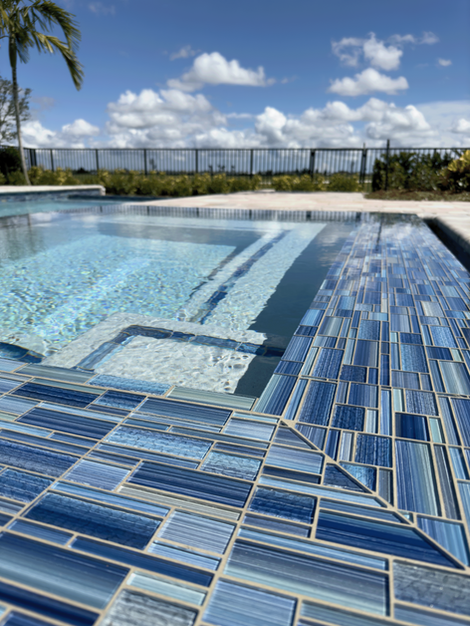 Sunlit water cascading down blue-tiled steps into a serene pool, framed by palm trees and a bright blue sky.