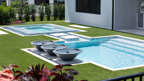 A modern backyard pool with water bowls, a raised spa with mosaic tiles, and a minimalist step-stone path, surrounded by lush greenery.