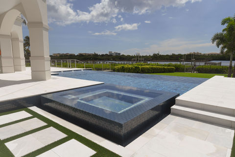 A modern waterfront pool with a raised spa, infinity-edge, and white stone patio, overlooking a waterway with tropical greenery.